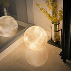 A lit Clérachic moon lamp on a glossy floor, with a second sphere reflected beside it and a vase of yellow blooms.