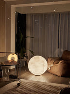 Clérachic moon sphere on a wooden floor, lit softly beside a beanbag chair and a metal trolley, with sheer curtains behind.