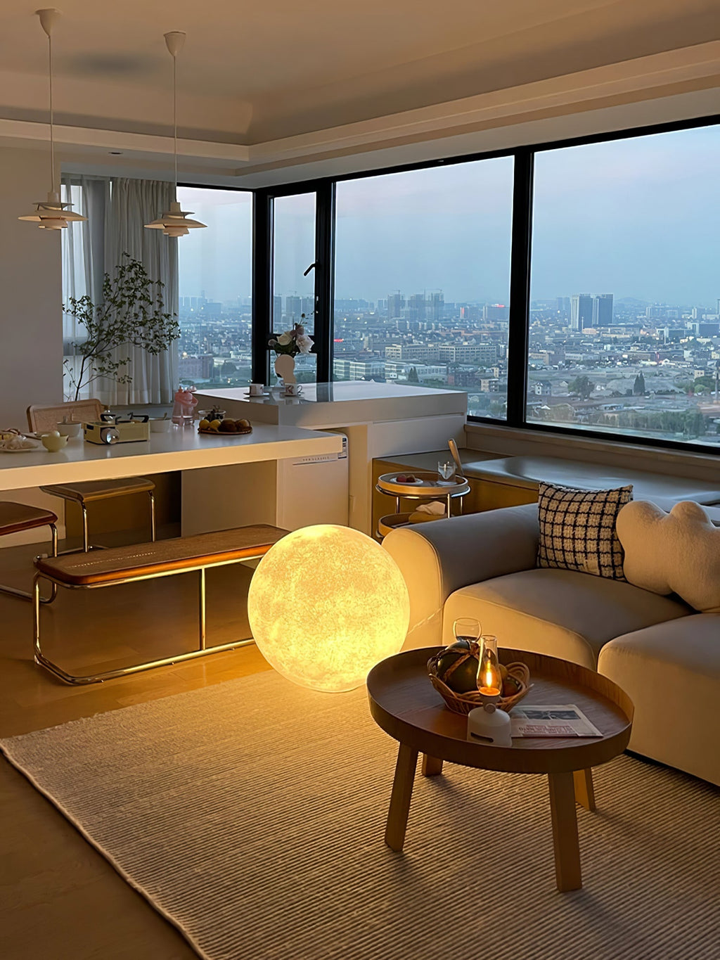 A small golden-lit Clérachic moon lamp on the floor of an open-plan room with a grey sofa, a round wooden table and city views.