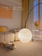 This Clérachic moon lamp is lit beside a cat on a woven carpet, with a cream sofa and an arched lamp in a pastel corner.