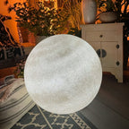Clérachic moon sphere with a soft grey-white surface, lit beside a white cabinet and a patterned rug in a plant-filled room.