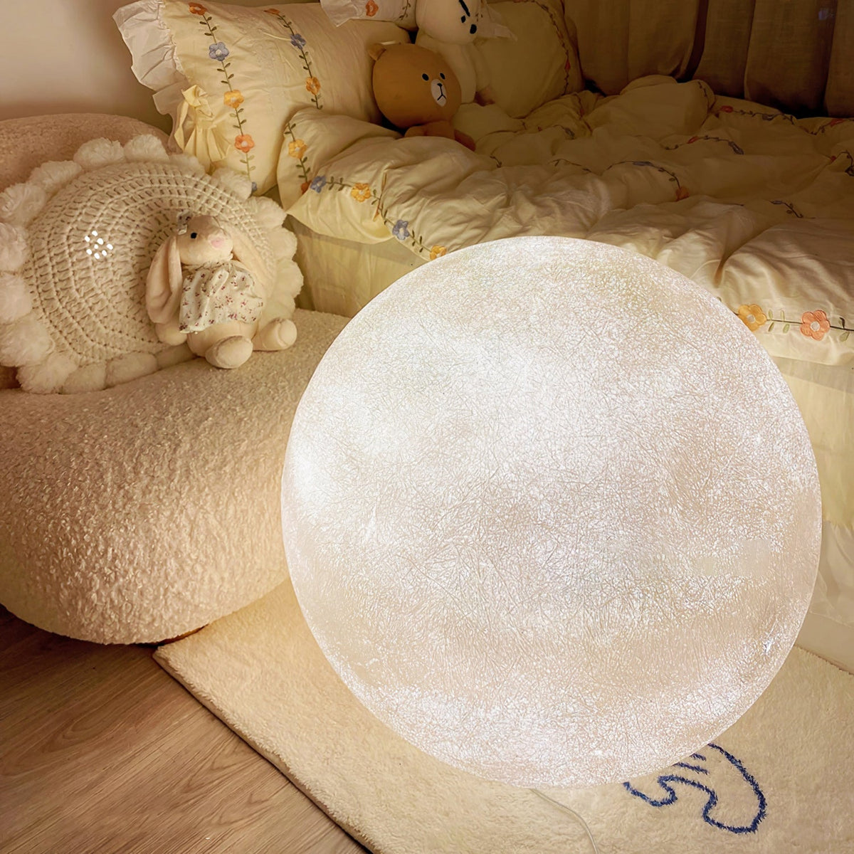 Close view of a bright white Clérachic moon sphere resting on a rug beside a boucle cushion, with soft toys and bedding behind.