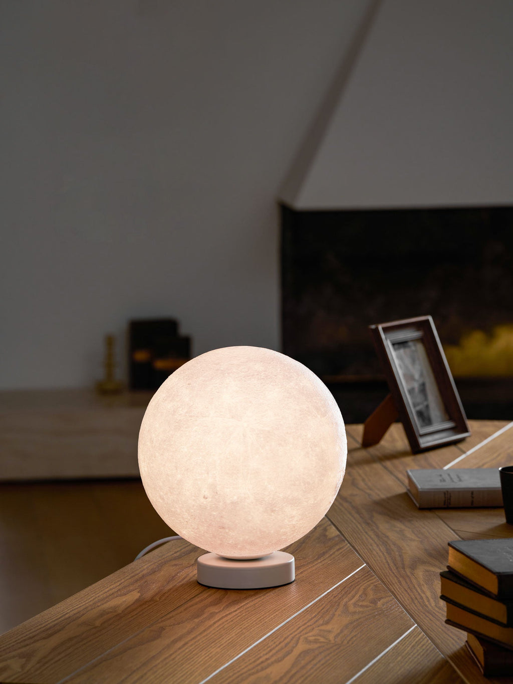 This Clérachic moon table lamp glows pink-white on a round white base, standing on a wooden desk with a photo frame and books.