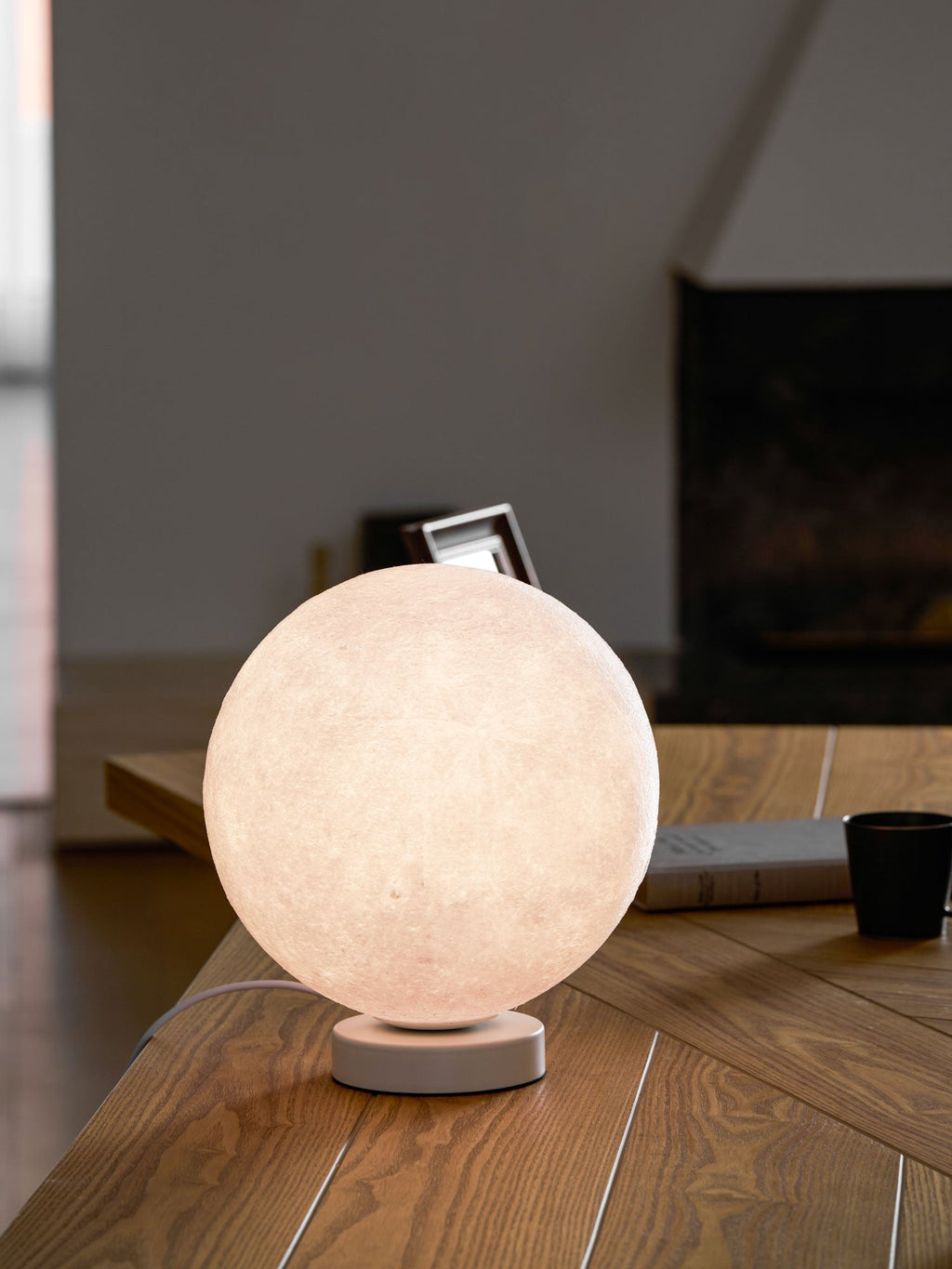 A closer view of the lit Clérachic moon table lamp on a wooden desk, its cratered surface glowing warm against a blurred dark wall.