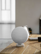 Unlit Clérachic moon table lamp on a white base, showing its pale, cratered grey surface, with a photo frame on a wooden desk.