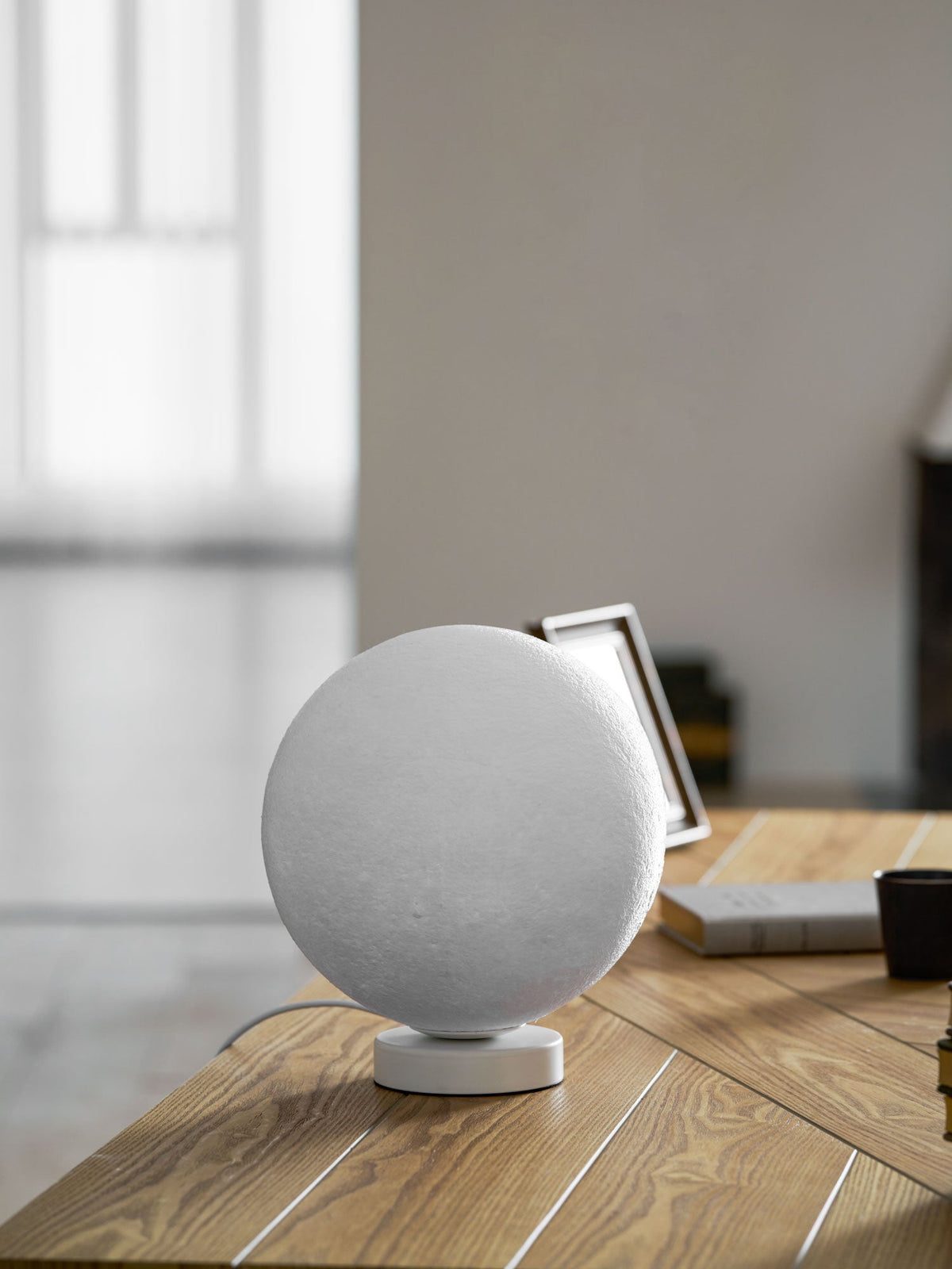 Unlit Clérachic moon table lamp on a white base, showing its pale, cratered grey surface, with a photo frame on a wooden desk.