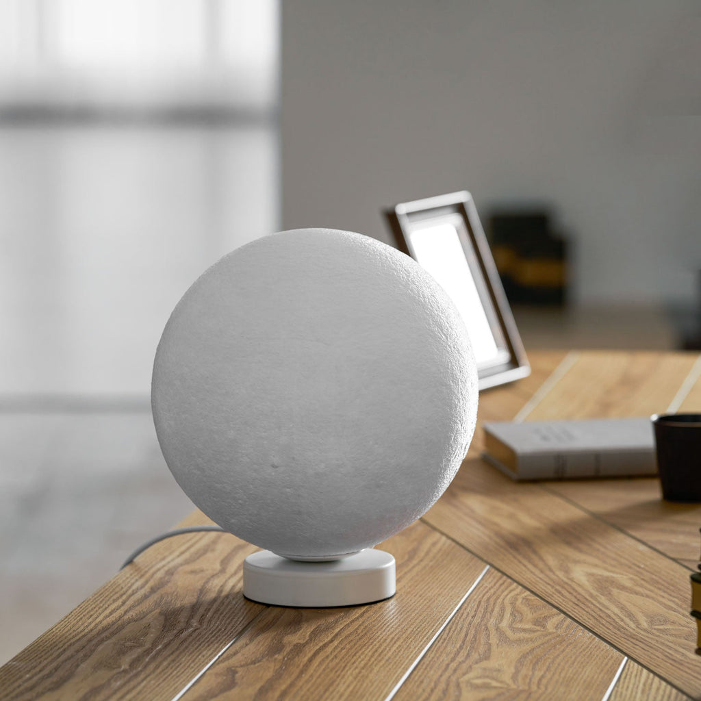 Medium-sized Clérachic moon table lamp, switched off, on its round white base on a wooden desk beside a stack of books.
