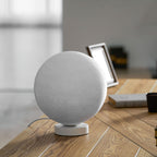 Medium-sized Clérachic moon table lamp, switched off, on its round white base on a wooden desk beside a stack of books.