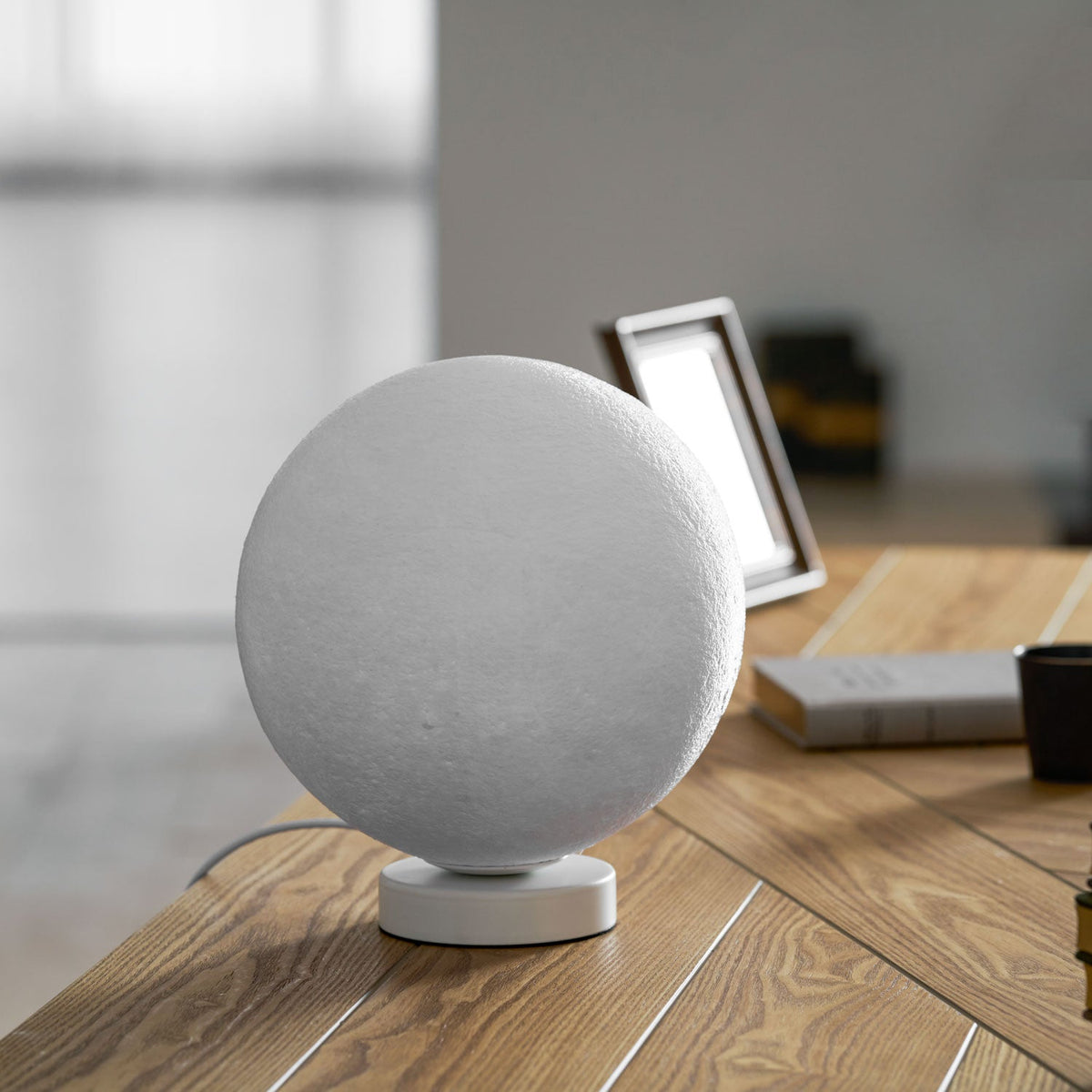 Medium-sized Clérachic moon table lamp, switched off, on its round white base on a wooden desk beside a stack of books.