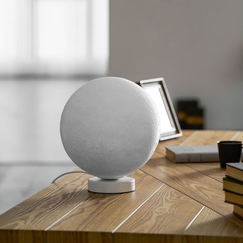Angled view of the unlit Clérachic moon table lamp on a wooden desk, with a photo frame and books behind it.