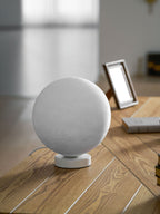 Small unlit Clérachic moon table lamp on its white base, standing on a wood-plank desk next to a hardback book.