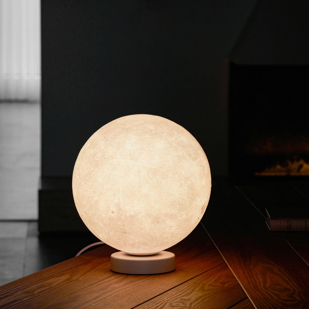 Lit at night, this Clérachic moon table lamp glows warm on a wooden desk in front of a dark fireplace.