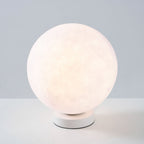 Lit Clérachic moon table lamp with a pink-white glow on a round white base, set on a pale surface against a blue-grey wall.