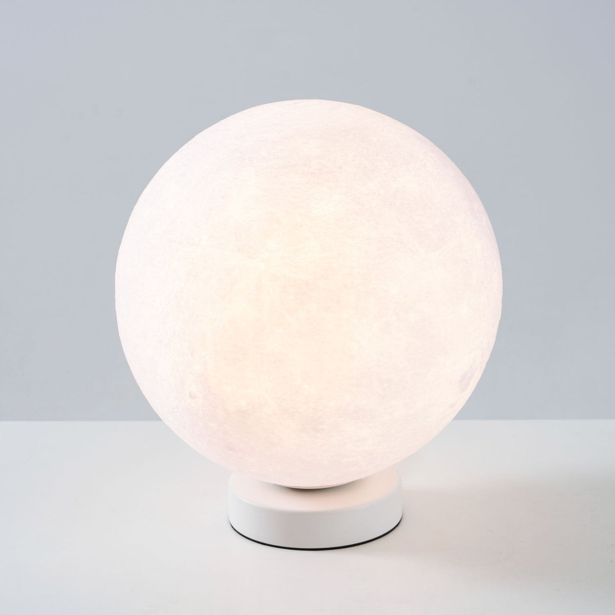 Lit Clérachic moon table lamp with a pink-white glow on a round white base, set on a pale surface against a blue-grey wall.