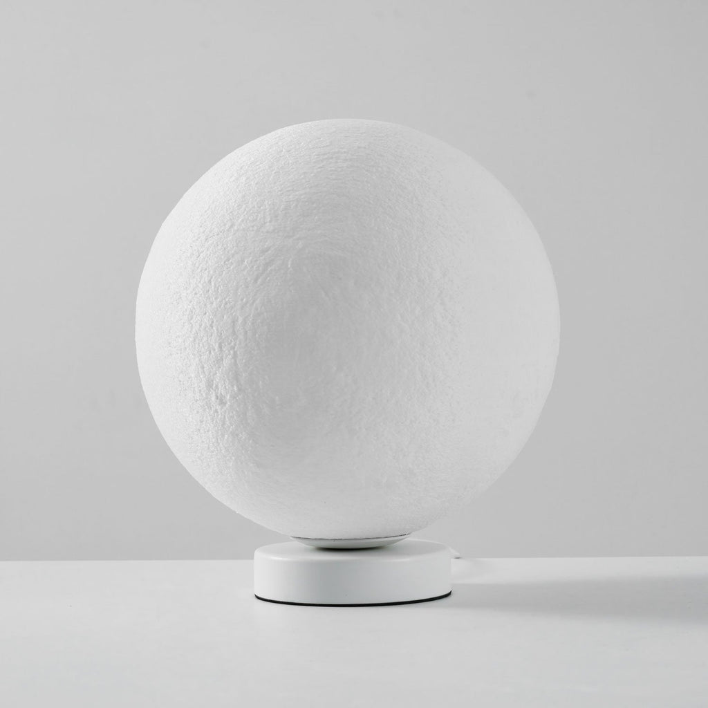 Unlit Clérachic moon table lamp, a white cratered sphere on a round white base, on a pale grey surface.