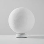 Unlit Clérachic moon table lamp, a white cratered sphere on a round white base, on a pale grey surface.