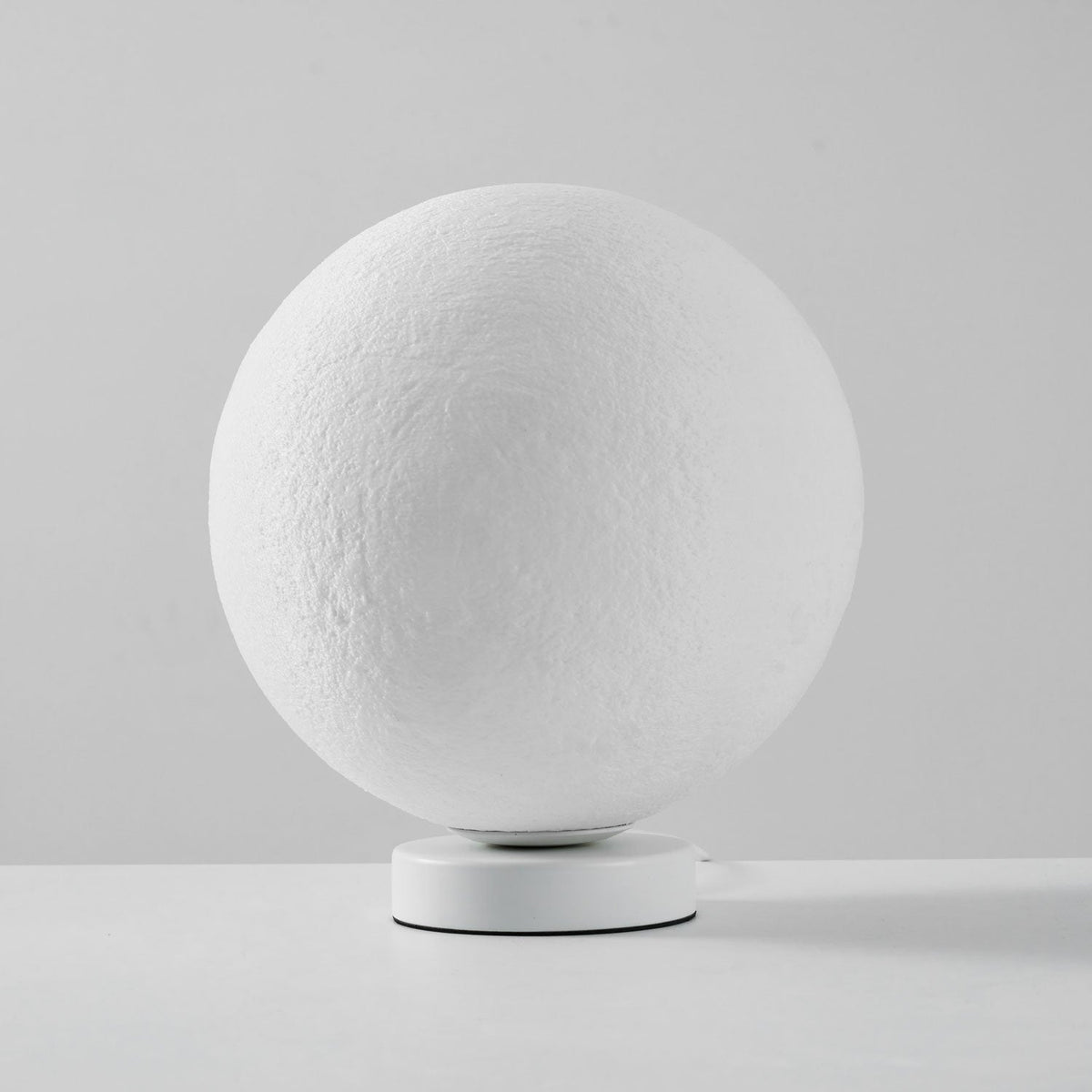 Unlit Clérachic moon table lamp, a white cratered sphere on a round white base, on a pale grey surface.