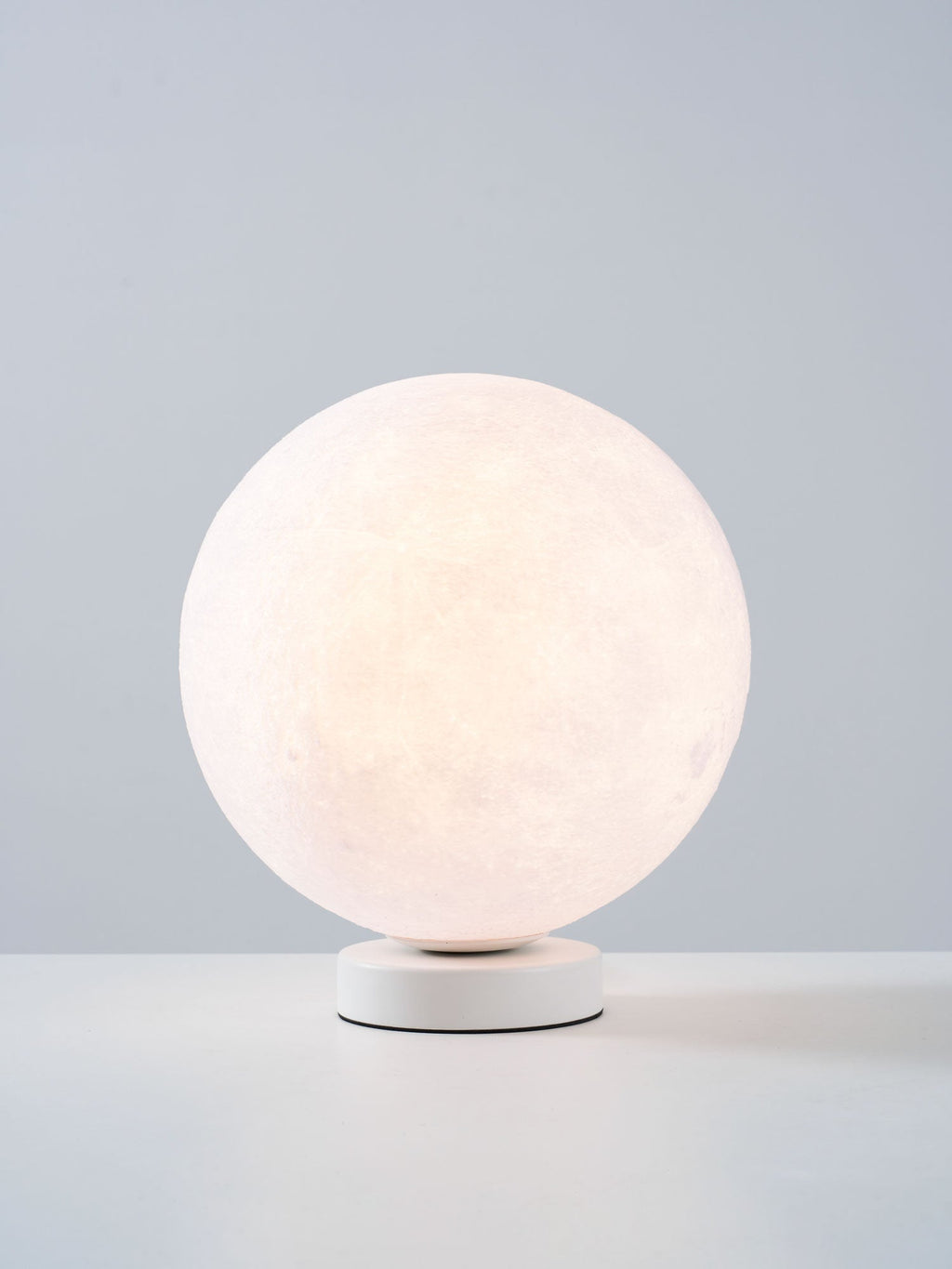 A smaller view of the lit Clérachic moon table lamp, glowing soft white on its base against a blue-grey backdrop.