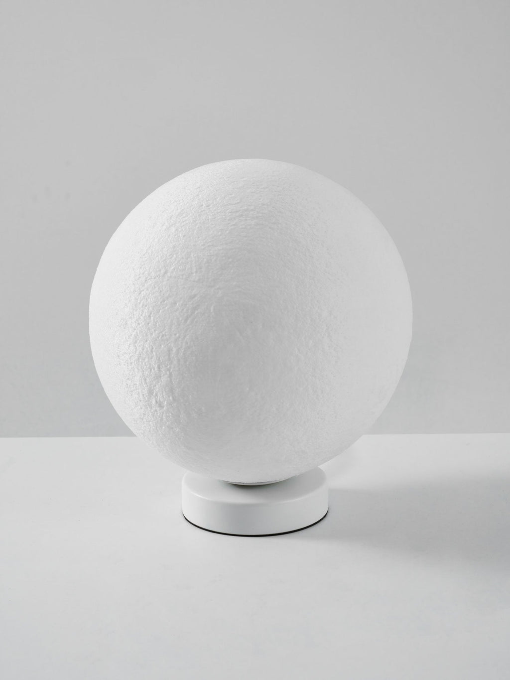 Side view of the unlit Clérachic moon lamp, its white sphere tilted on a low round base against a grey wall.