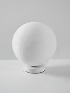 Side view of the unlit Clérachic moon lamp, its white sphere tilted on a low round base against a grey wall.