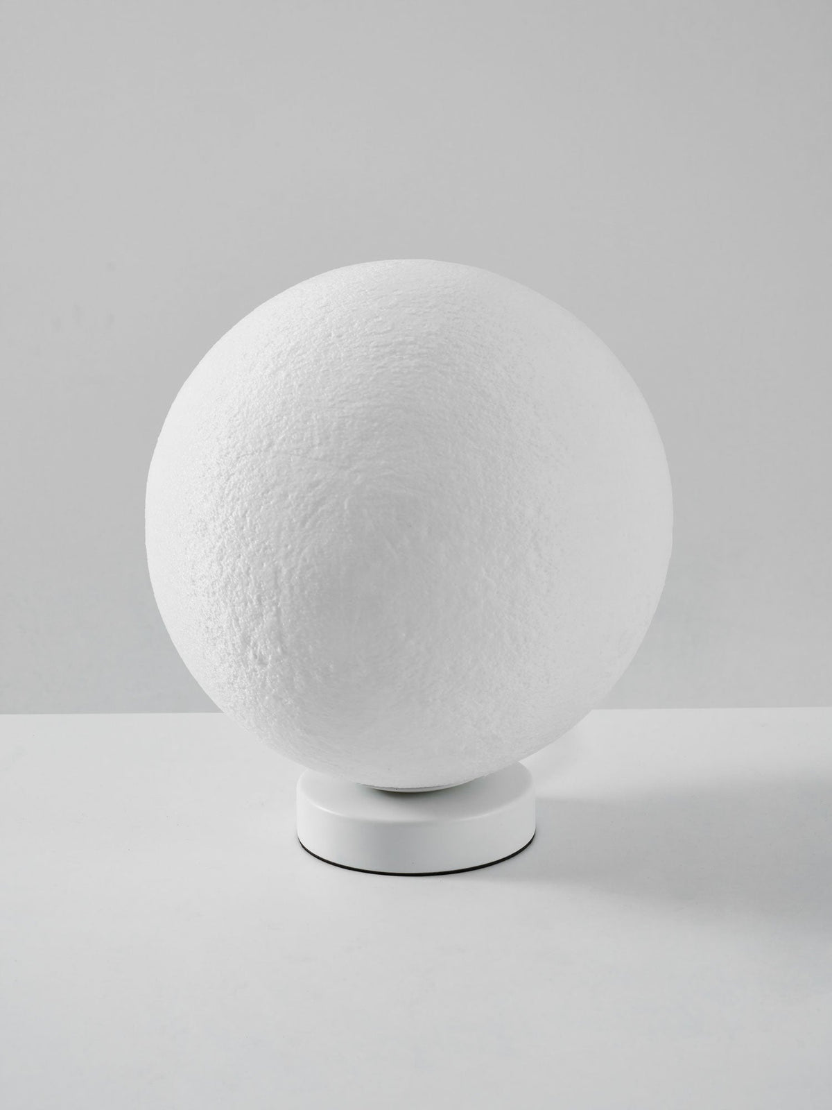 Side view of the unlit Clérachic moon lamp, its white sphere tilted on a low round base against a grey wall.