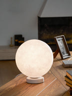 Clérachic moon table lamp glowing warm on a wooden desk, with a framed photo and a stack of books beside it.