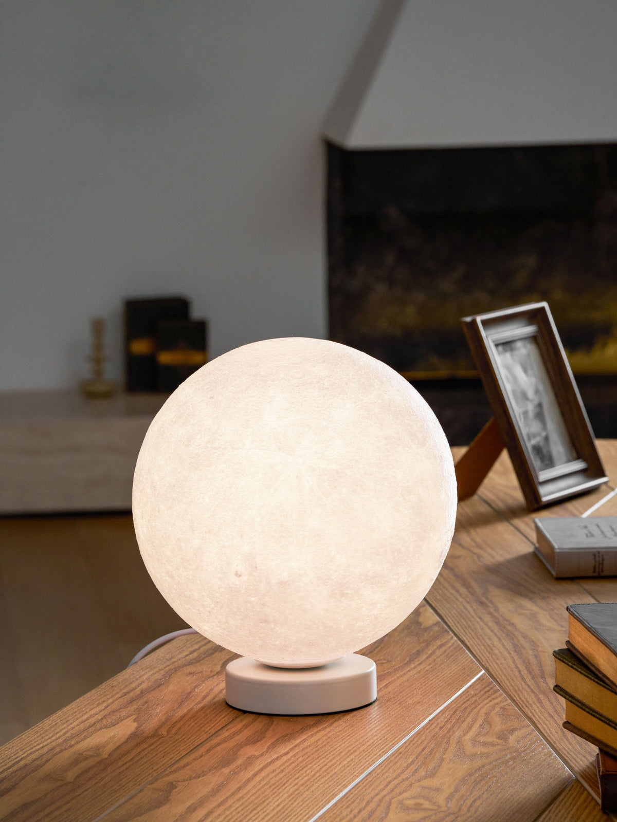 Clérachic moon table lamp glowing warm on a wooden desk, with a framed photo and a stack of books beside it.
