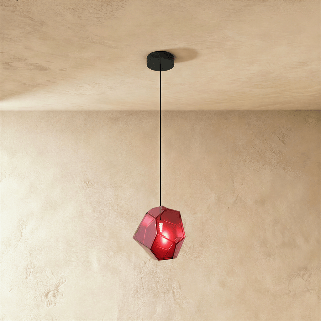 A Clérachic faceted glass pendant in deep rose red hangs on a black cord against a warm plastered ceiling corner.
