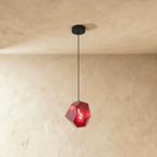 A Clérachic faceted glass pendant in deep rose red hangs on a black cord against a warm plastered ceiling corner.