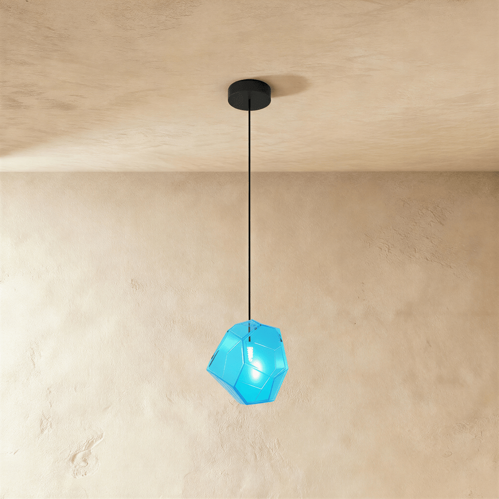 A Clérachic faceted glass pendant in bright blue hangs on a black cord against a warm plastered ceiling corner.