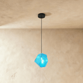 A Clérachic faceted glass pendant in bright blue hangs on a black cord against a warm plastered ceiling corner.