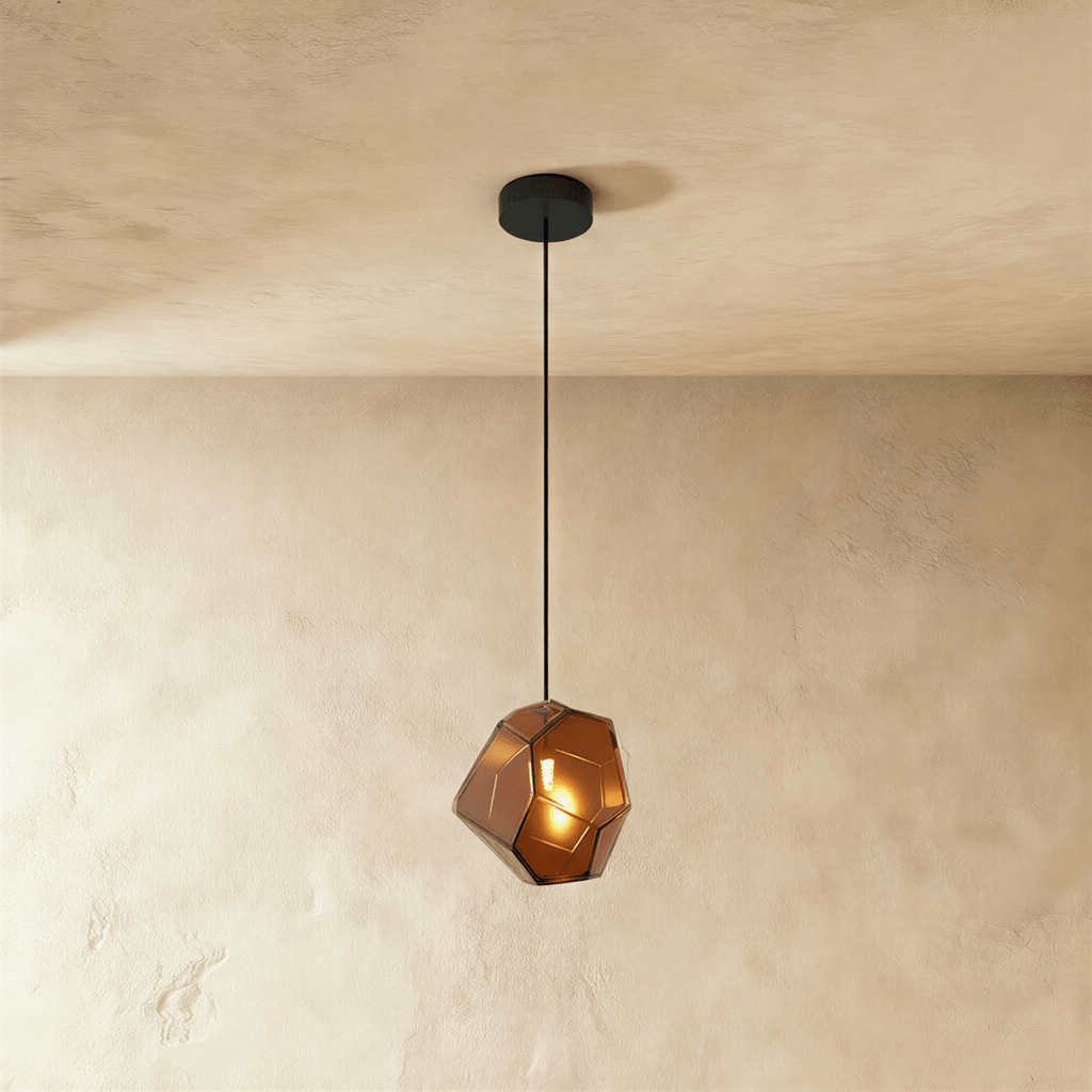 A Clérachic faceted glass pendant in warm brown hangs on a black cord against a plastered ceiling corner.