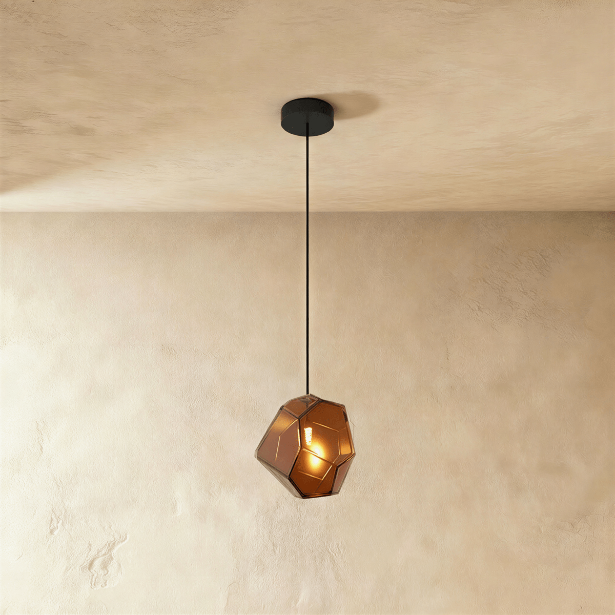 A Clérachic faceted glass pendant in warm brown hangs on a black cord against a plastered ceiling corner.
