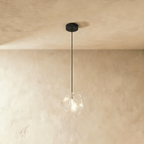 A Clérachic faceted glass pendant in clear glass hangs on a black cord against a warm plastered ceiling corner.