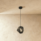 A Clérachic faceted glass pendant in smoked light gray hangs on a black cord against a plastered ceiling corner.