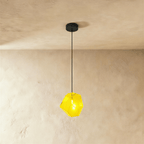 A Clérachic faceted glass pendant in bright yellow hangs on a black cord against a warm plastered ceiling corner.