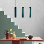 Three green Clérachic glass tube pendants hung in a row above a coral dining table beside a striped green staircase.