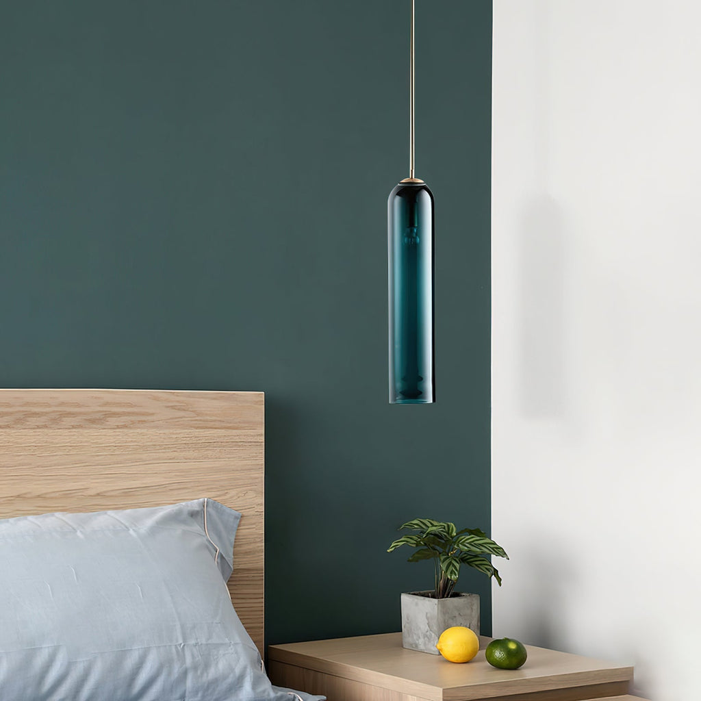 A green Clérachic glass tube pendant lit above a wooden nightstand with a potted plant, against a dark teal wall.