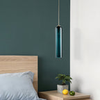 A green Clérachic glass tube pendant lit above a wooden nightstand with a potted plant, against a dark teal wall.