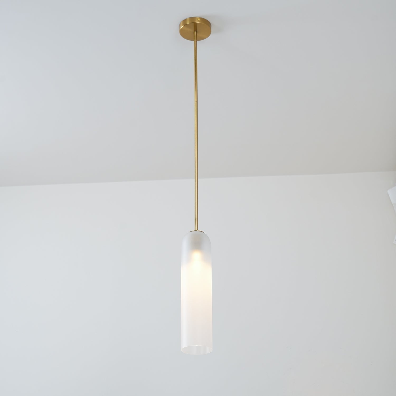 A frosted white Clérachic tube pendant lit on a brass rod, hung in a bright room with a sheer curtain at the edge.