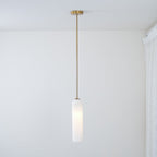 The frosted white Clérachic pendant lit on a brass rod, its glass catching light near a curtained window corner.