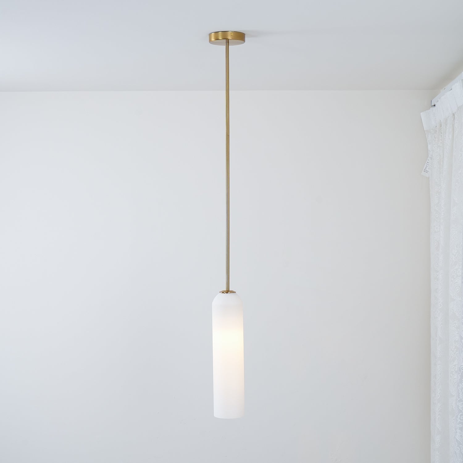 The frosted white Clérachic pendant lit on a brass rod, its glass catching light near a curtained window corner.