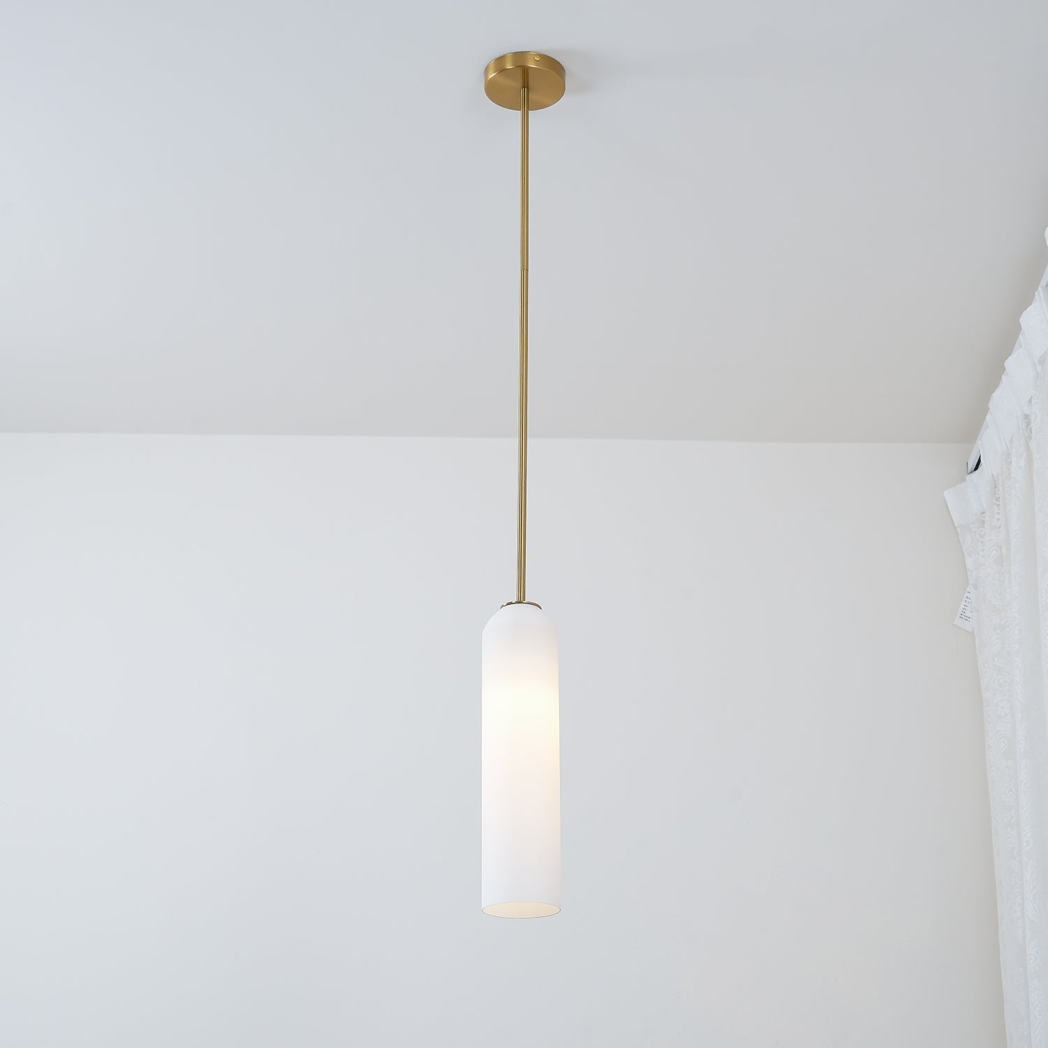 A frosted white Clérachic tube pendant lit on a brass rod, hung close to a lace curtain in a sunlit room.
