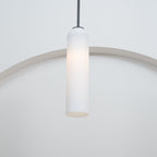A frosted white Clérachic tube pendant lit on a brass rod, hung in a plain room with a curtain at the frame's edge.