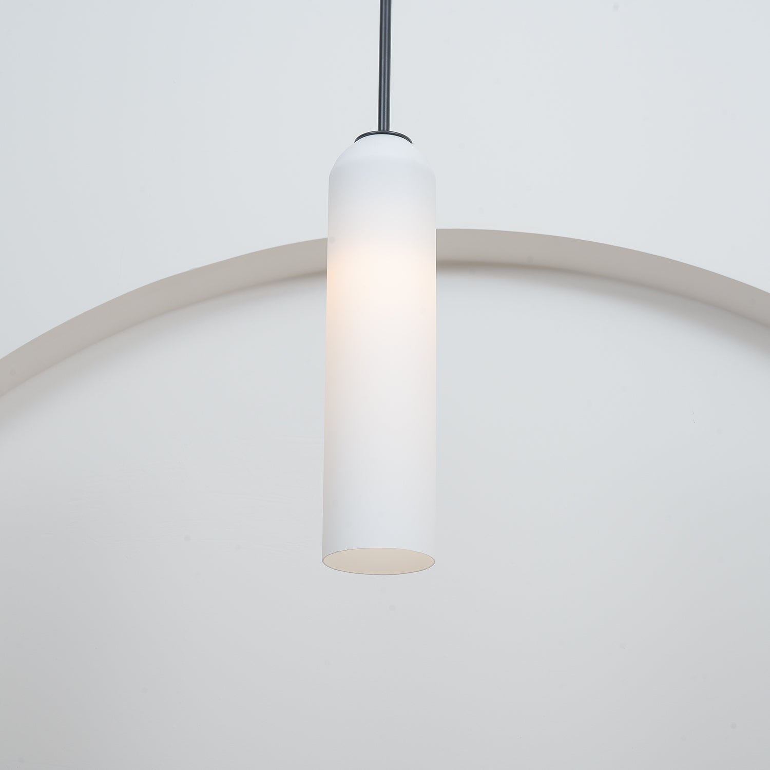 A frosted white Clérachic tube pendant lit on a brass rod, hung in a plain room with a curtain at the frame's edge.