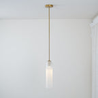 The frosted white Clérachic pendant on a brass rod, lit, its long stem reaching down near a curtained window.