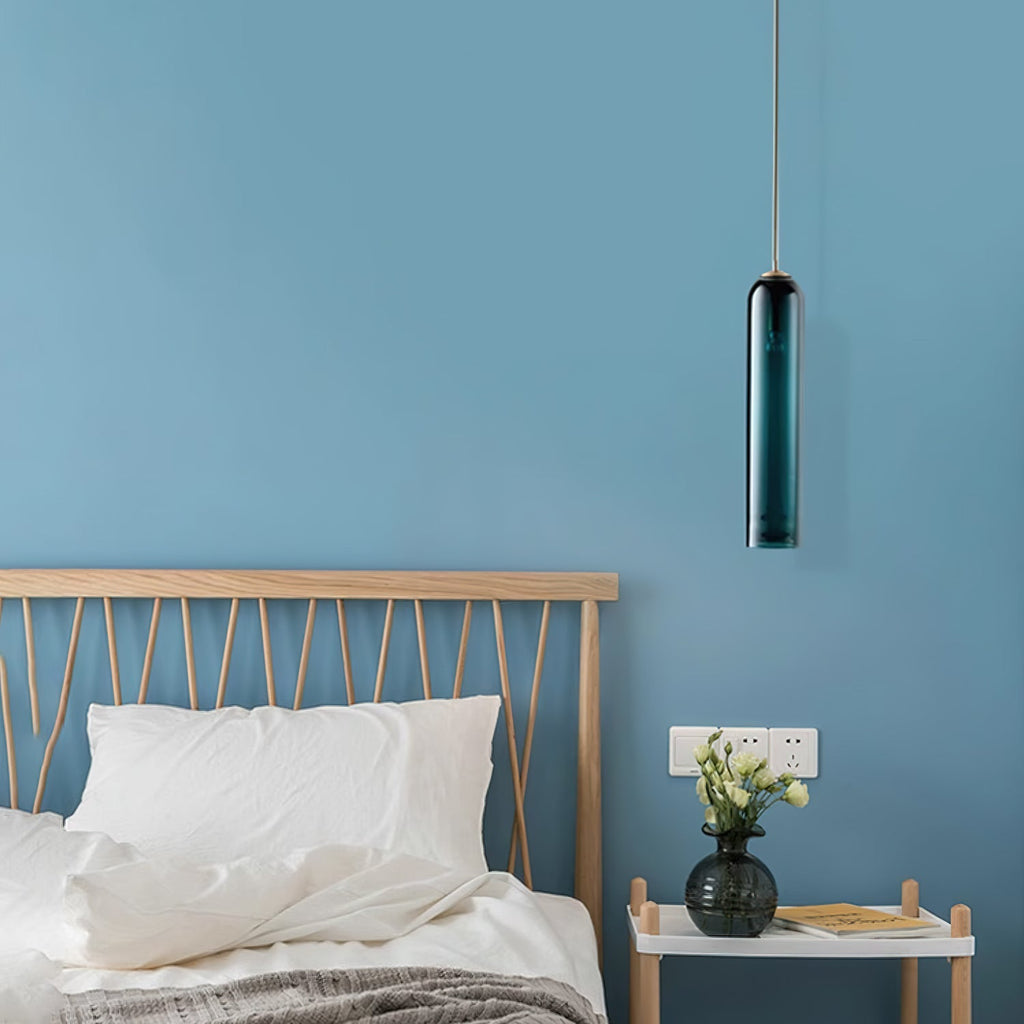 A green Clérachic glass tube pendant lit above a bedside table with a vase and books, against a blue wall.