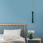 A green Clérachic glass tube pendant lit above a bedside table with a vase and books, against a blue wall.