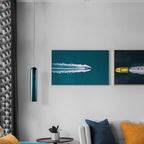 A green Clérachic glass tube pendant lit in a living room, beside framed aerial photography and a grey sofa.