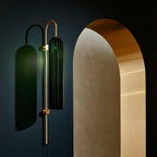 Clérachic mouth-blown glass wall sconce in green against a navy wall, throwing a tall shadow next to a brass and stone arch.